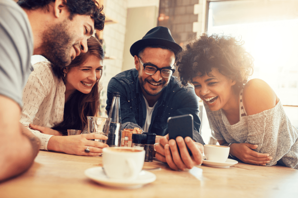 Portrait of cheerful young friends looking at smart phone while sitting in cafe. Mixed race people sitting at a table in restaurant using mobile phone.